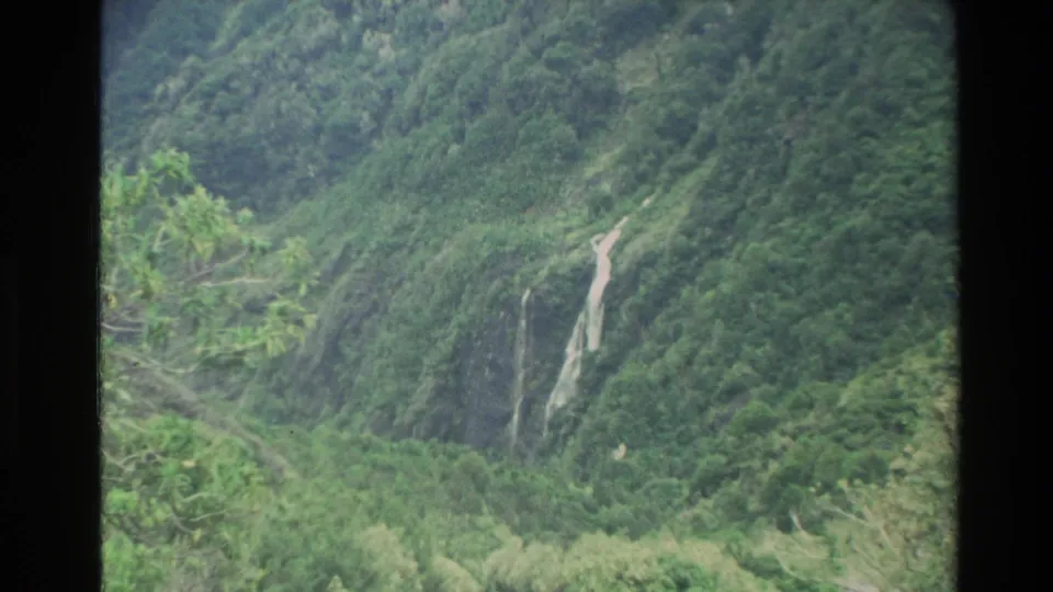 1982 New Zealand home-movie frame focused on a waterfall cutting down a steep, forested valley slope with soft 8mm texture.