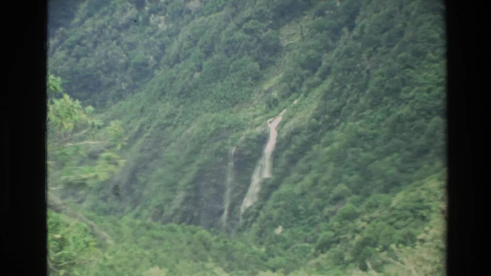 1982 New Zealand 8mm frame showing a lush mountain valley slope with a small white waterfall descending through greenery.