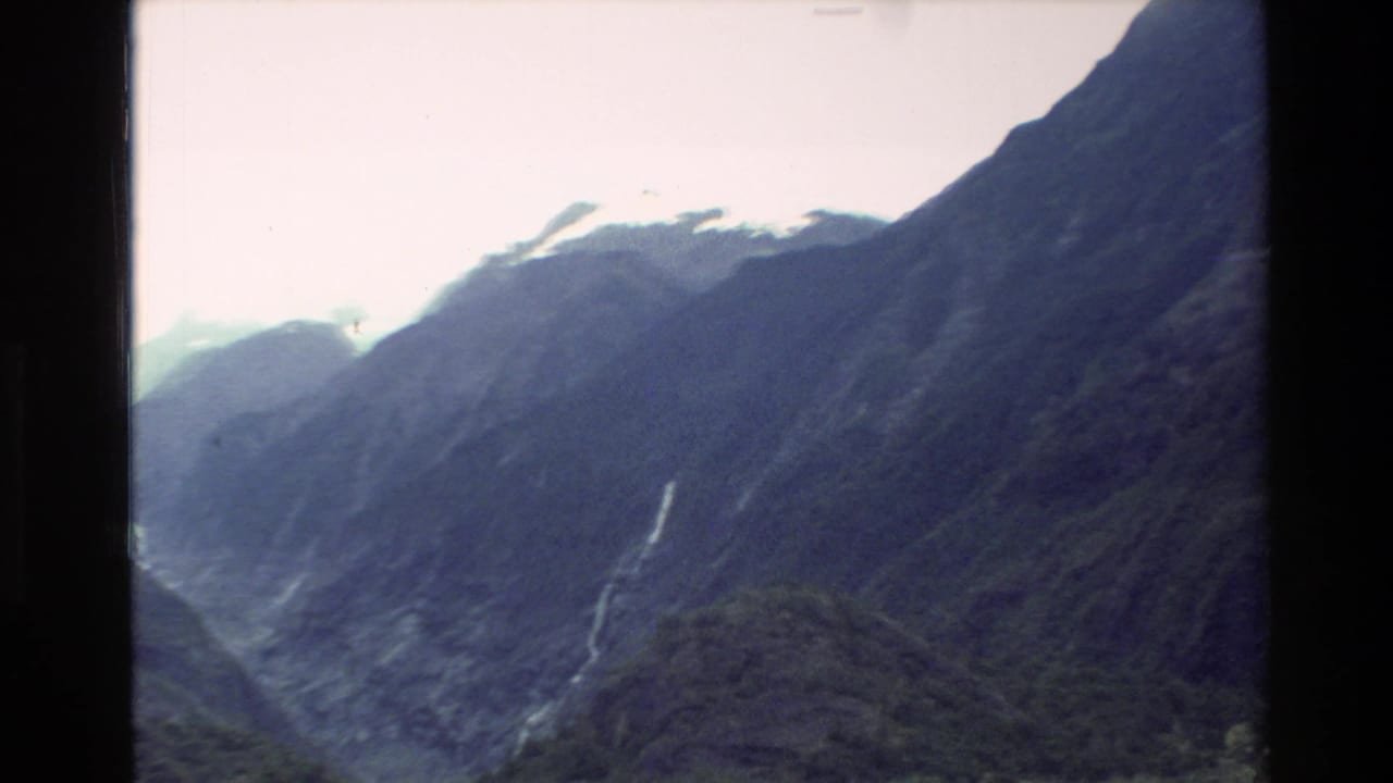 1982 New Zealand 8mm frame of dark alpine mountains, distant snow or ice, and a slim waterfall under overcast sky.