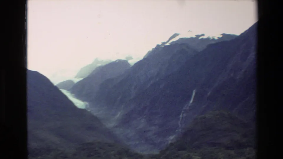 1982 New Zealand 8mm frame looking across dark alpine slopes toward pale snow or ice and a narrow waterfall.