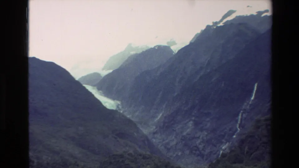 1982 New Zealand 8mm frame of a broad alpine valley with distant snow or ice, dark ridges, and thin waterfalls.