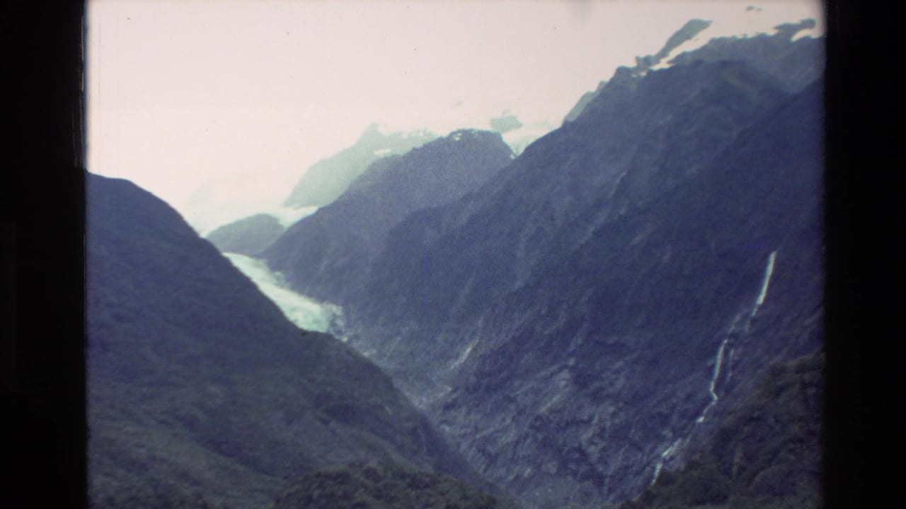 1982 New Zealand 8mm frame showing a rugged mountain valley with a pale glacier or snowfield and a thin waterfall on a dark slope.