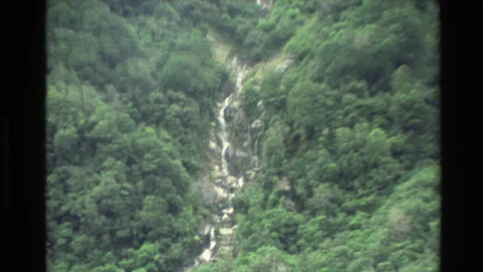 1982 New Zealand 8mm frame with handheld softness, showing a white waterfall channel through dense green forest.