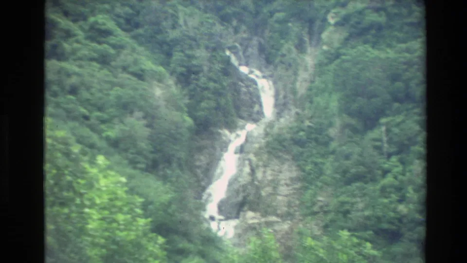 Vintage 8mm view of a narrow waterfall dropping through dense green New Zealand mountain forest.
