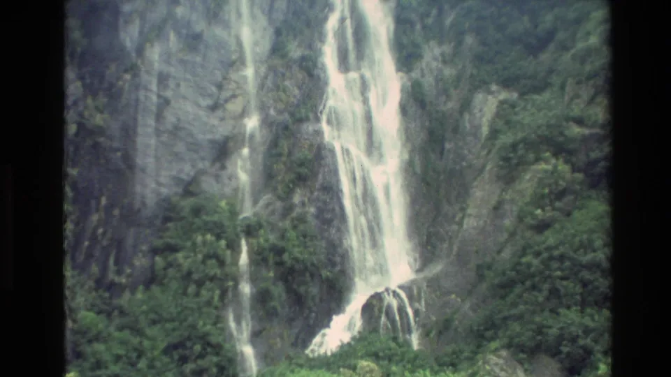 1982 New Zealand 8mm frame showing multiple waterfall ribbons over a rugged cliff with dense greenery below.