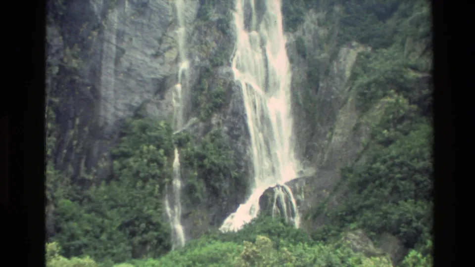 1982 New Zealand 8mm frame with a broad waterfall descending a dark cliff among trees and vegetation.