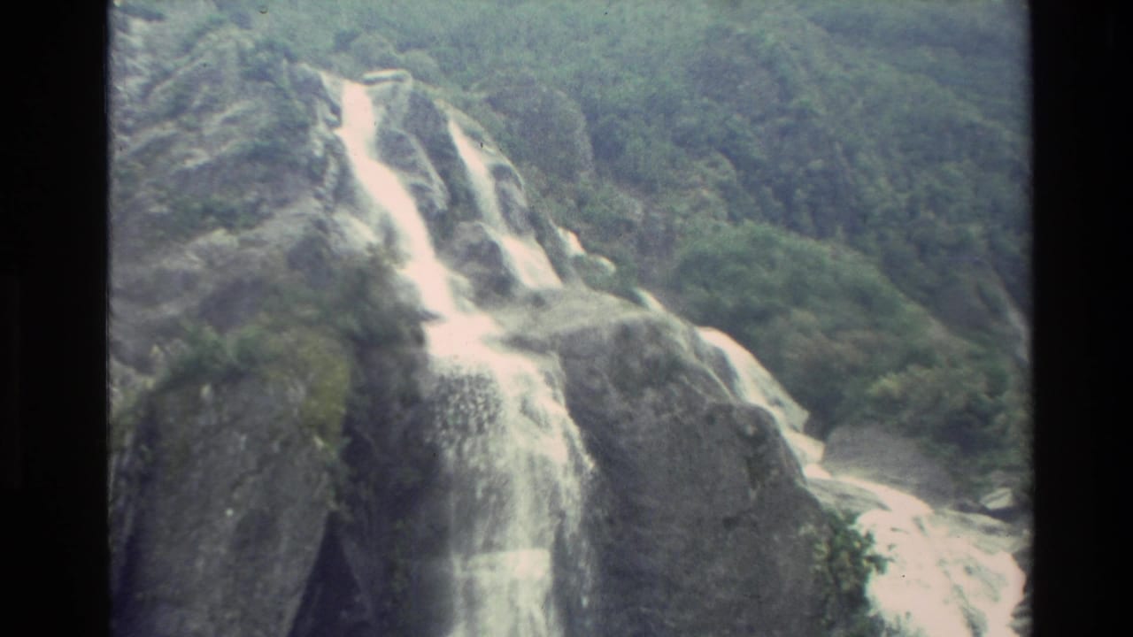 1982 New Zealand 8mm frame showing a waterfall spilling over rugged rock ledges amid dense green mountain vegetation.