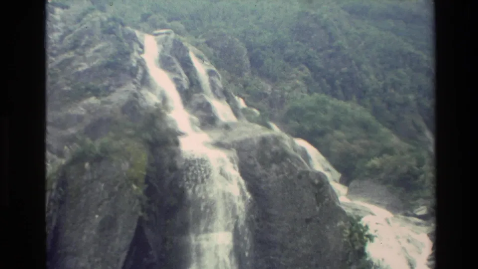 1982 New Zealand 8mm frame showing a waterfall spilling over rugged rock ledges amid dense green mountain vegetation.