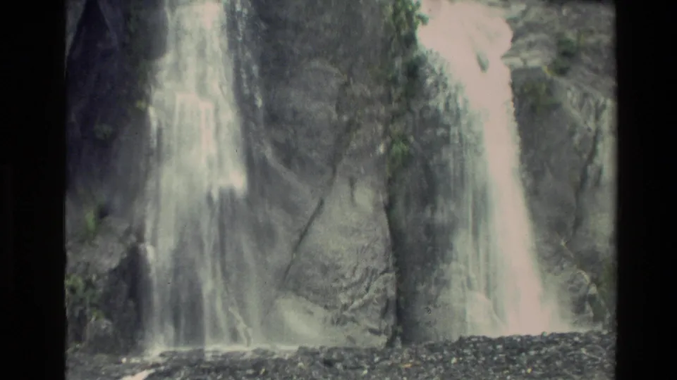 1982 New Zealand home-movie frame showing water dropping across a broad rocky surface with pale spray and greenery nearby.