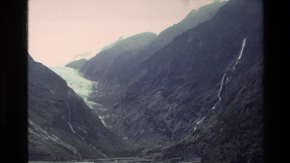 1982 New Zealand 8mm frame showing a wide mountain valley with dark slopes, a pale distant water or ice feature, and narrow cascades.