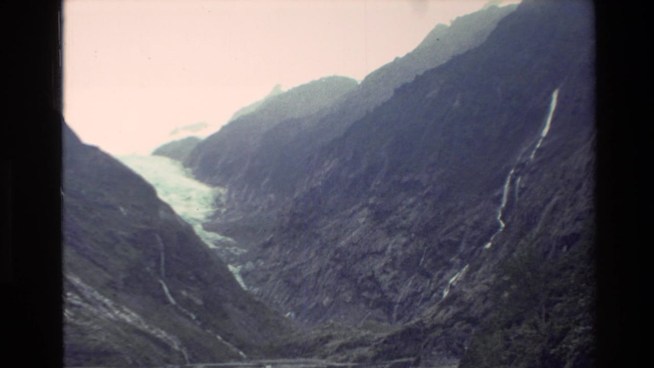 1982 New Zealand 8mm frame showing a glacier descending between steep rocky mountains with narrow waterfalls on the valley sides.