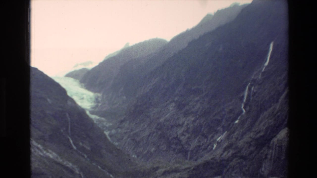 Glacier and waterfalls viewed across dark mountain slopes in a narrow valley.