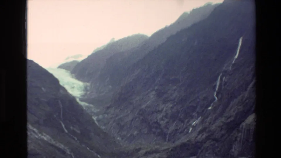 1982 New Zealand travel-film frame with a distant glacier, shadowed rocky slopes, and several pale waterfall streaks.
