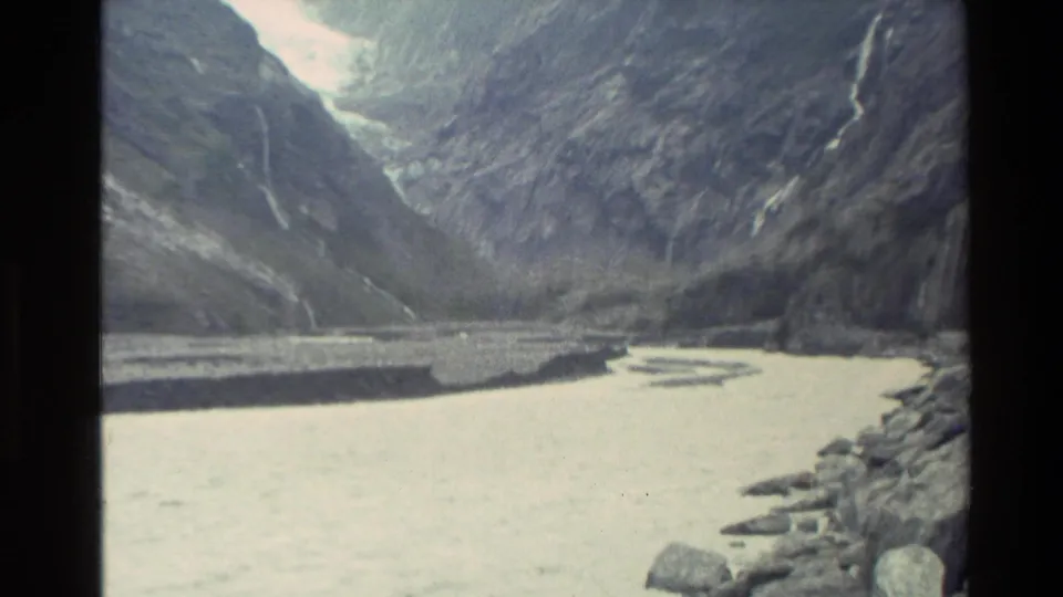 1982 New Zealand 8mm frame looking down toward a pale glacial river beneath steep mountains and waterfall lines.