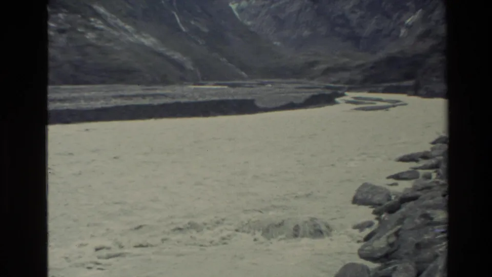 1982 New Zealand home-movie frame of a pale turbulent river crossing a rocky alpine valley with waterfalls on dark slopes.