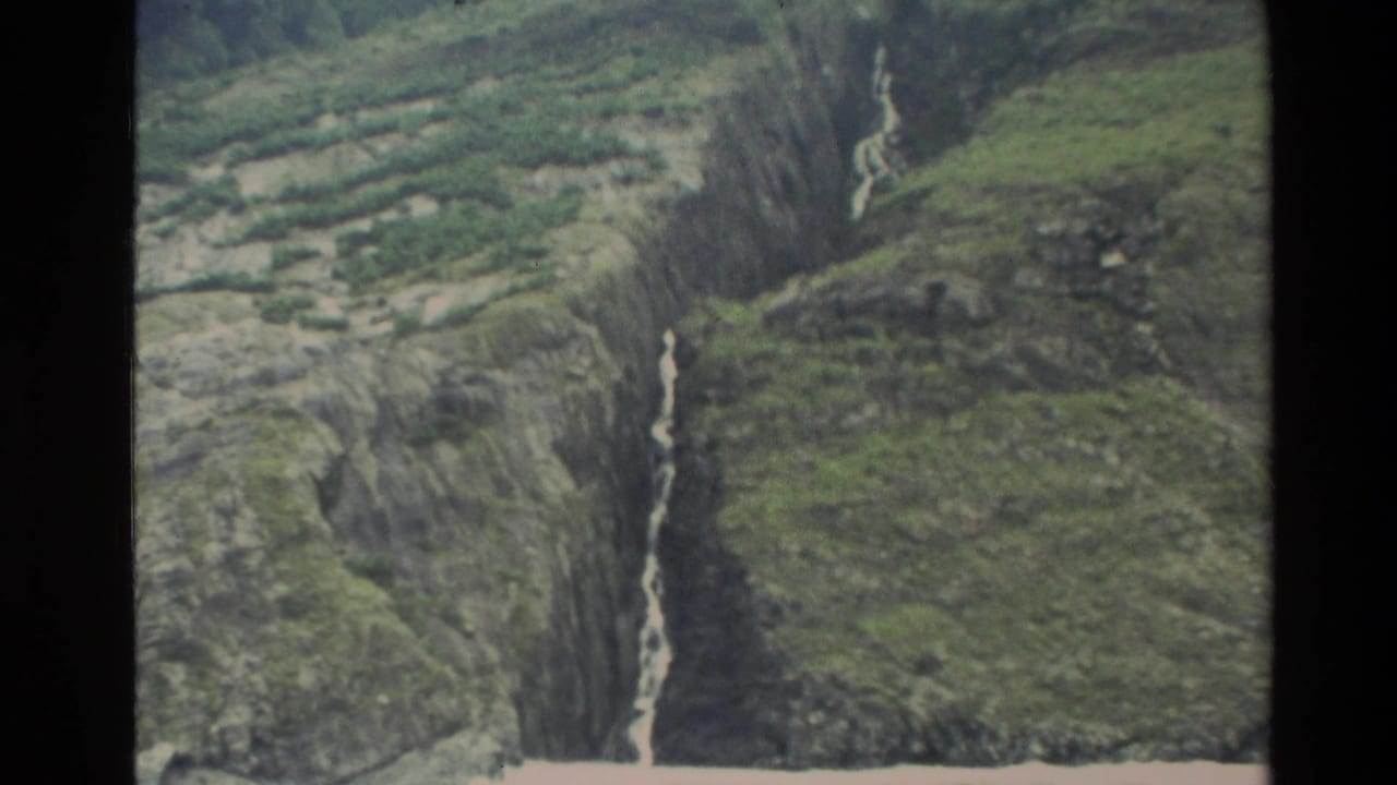 1982 New Zealand 8mm frame showing a steep green valley wall with a thin waterfall descending through dark rock and vegetation.