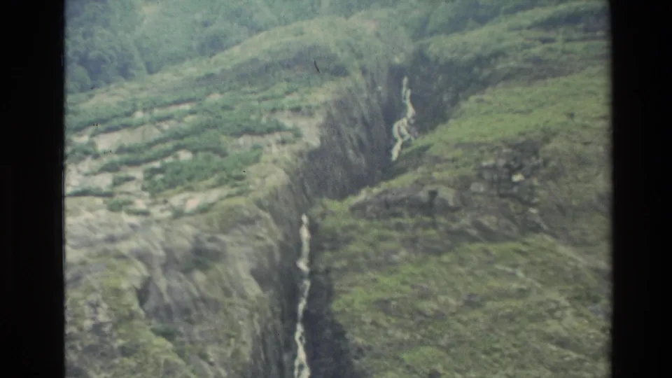 1982 New Zealand home-movie frame of green mountain slopes and pale watercourses descending through a rugged ravine.