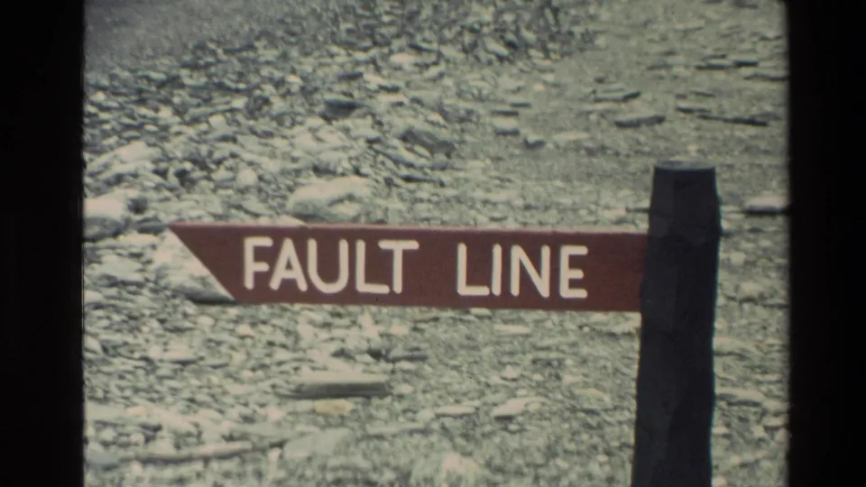 1982 New Zealand 8mm frame with a red FAULT LINE sign in front of a pale rocky riverbed and rugged valley floor.