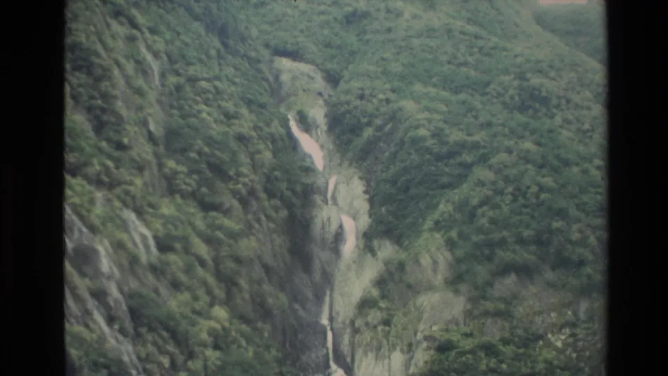 1982 New Zealand frame showing a green gorge, pale watercourse, and narrow waterfall within steep mossy-looking slopes.