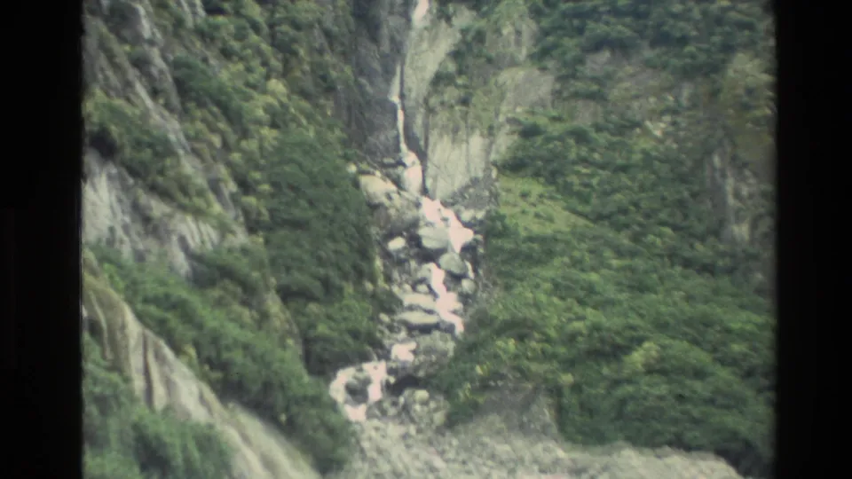 1982 New Zealand 8mm frame with a cascade flowing over boulders on a lush, steep mountain slope.