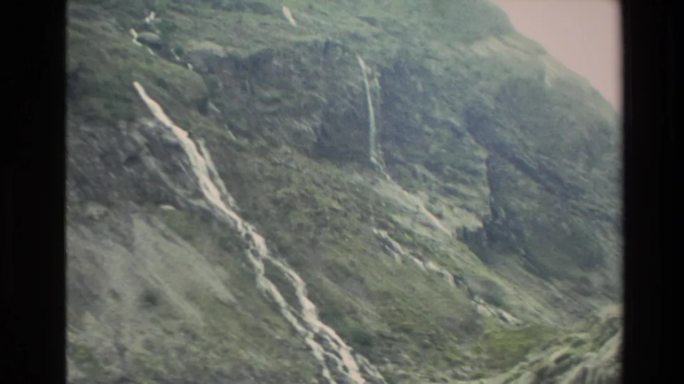 1982 New Zealand 8mm frame of a long white waterfall cutting diagonally down a rocky, vegetated mountainside.