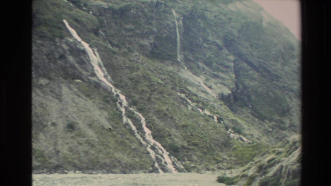 1982 New Zealand 8mm frame showing white waterfalls running down steep green cliffs in a mountain valley.