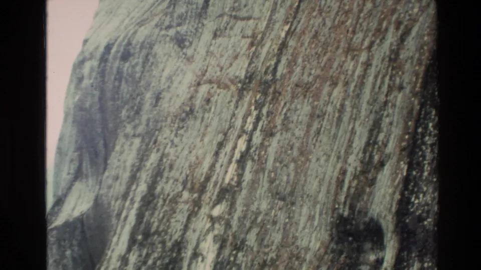 1982 New Zealand 8mm frame with a steep textured rock face filling the foreground and valley terrain beyond.