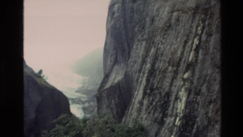 1982 New Zealand 8mm frame looking between dark rock walls toward a hazy mountain valley with vegetation below.