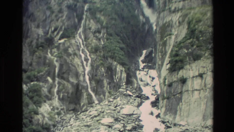 1982 New Zealand 8mm frame looking down the gorge to fast white water, loose rocks, and steep grey cliff walls with patches of greenery.