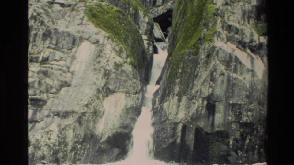 1982 New Zealand home-movie frame with a white waterfall cutting through gray rock and mossy mountain slopes.