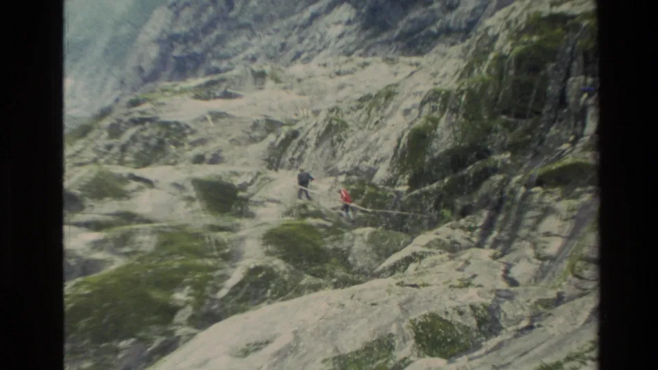 1982 New Zealand 8mm frame showing distant hikers, including a red jacket, moving across a rocky alpine trail.