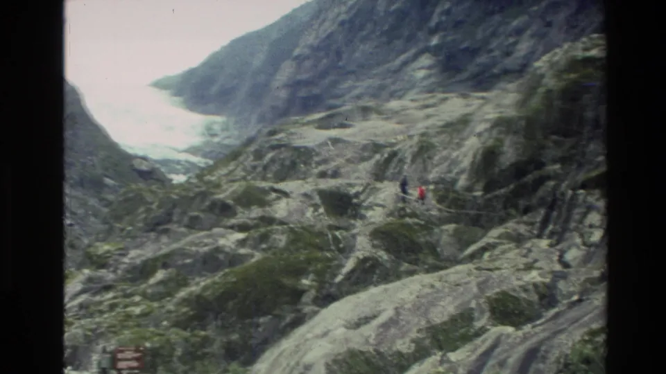 1982 New Zealand home-movie frame with small hikers on rough rock, backed by steep slopes and a bright distant valley or watercourse.