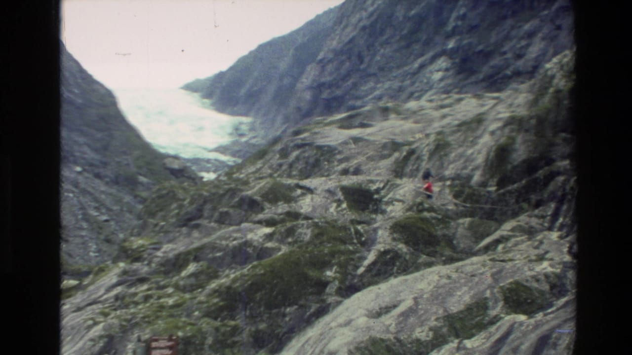 1982 New Zealand home-movie frame showing a rocky mountain valley, distant hikers, and pale glacier or water beyond the slope.