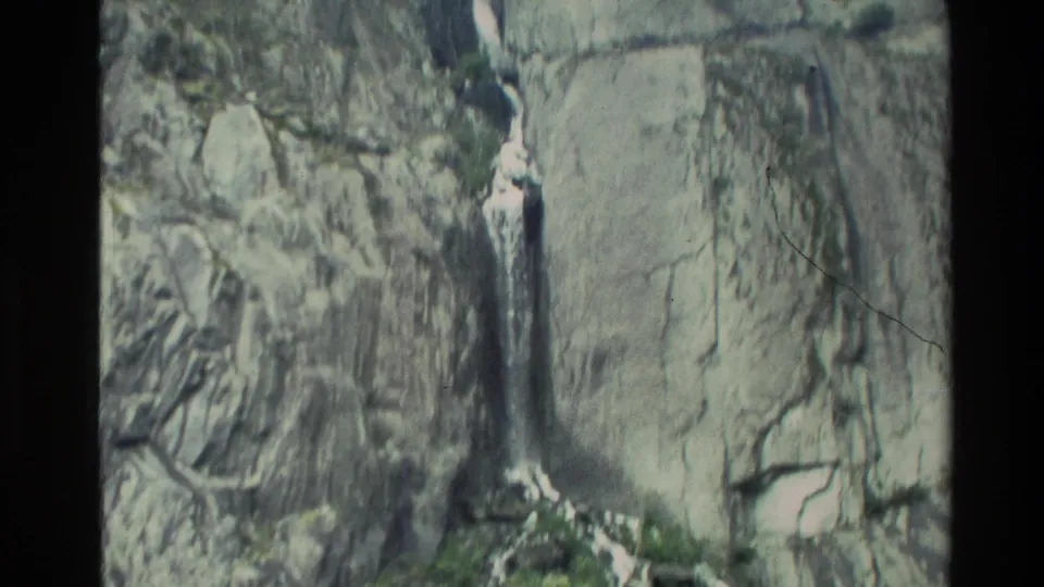 1982 New Zealand 8mm frame of a narrow waterfall descending a steep gray cliff with wet rock textures.