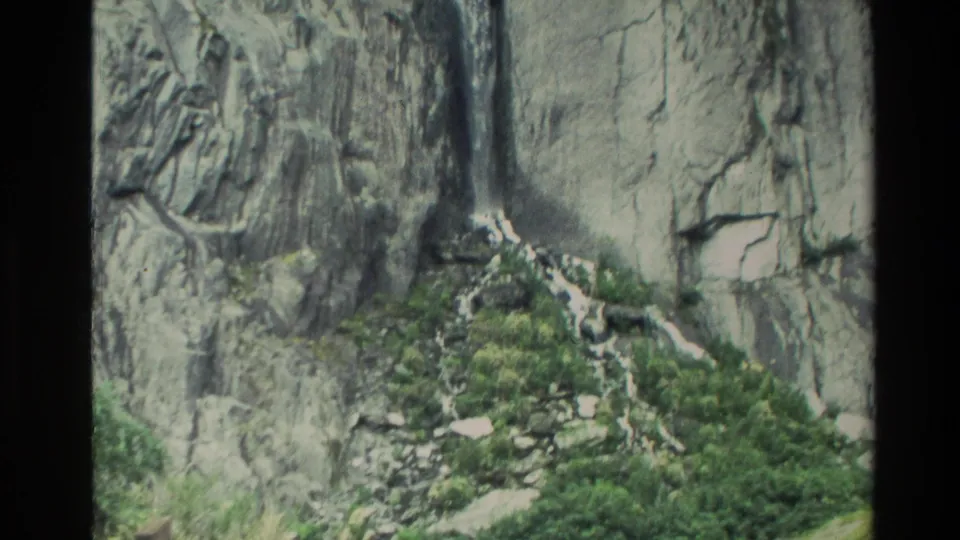 1982 New Zealand home-movie frame showing a cliff waterfall feeding a stream through rocks and green vegetation.