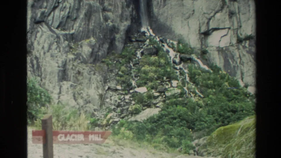 1982 New Zealand frame with a red Glacier Hill sign beside green scrub, boulders, and a cliff waterfall.