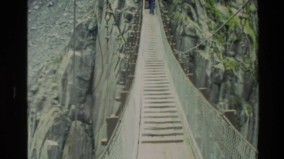 1982 New Zealand 8mm frame showing a wooden footbridge over a rocky gorge, with a distant person visible near the far end.
