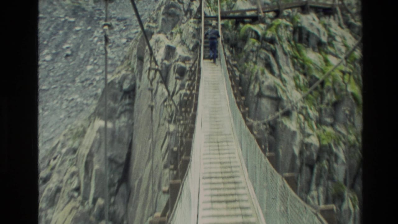 1982 New Zealand home-movie frame showing a man crossing a narrow suspension bridge between steep rock walls above rapids.