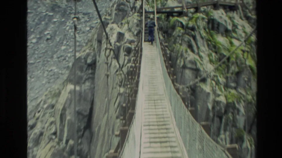 1982 New Zealand home-movie frame showing a man crossing a narrow suspension bridge between steep rock walls above rapids.