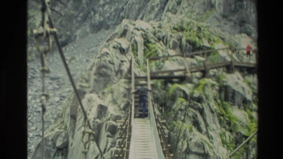 The bridge crossing continues in a rocky New Zealand gorge, with a distant visitor near timber steps and platforms.
