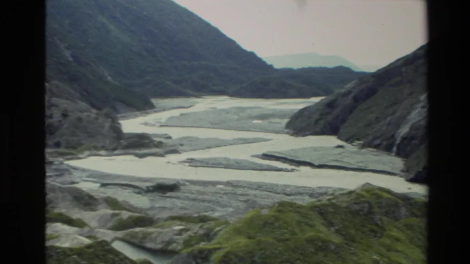1982 New Zealand 8mm frame looking over pale river flats and steep mountain slopes from an elevated viewpoint.