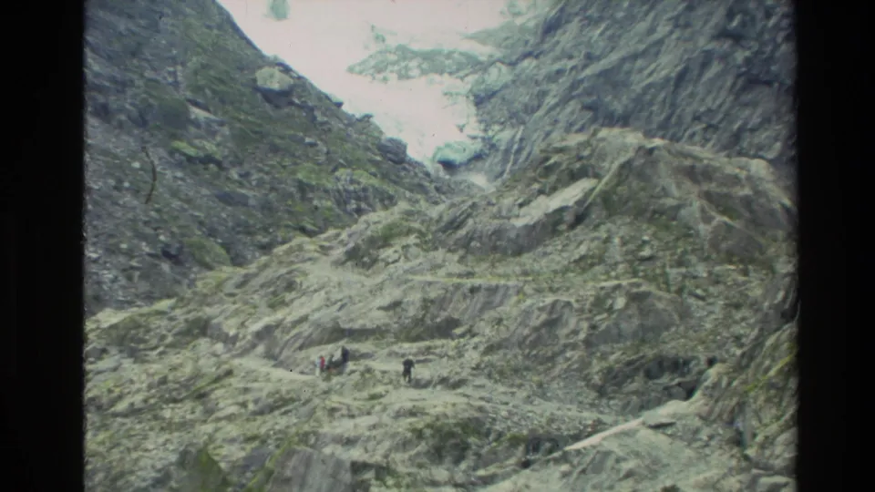 1982 New Zealand 8mm frame of a rocky glacier valley with tiny walkers visible below the ice and slopes.