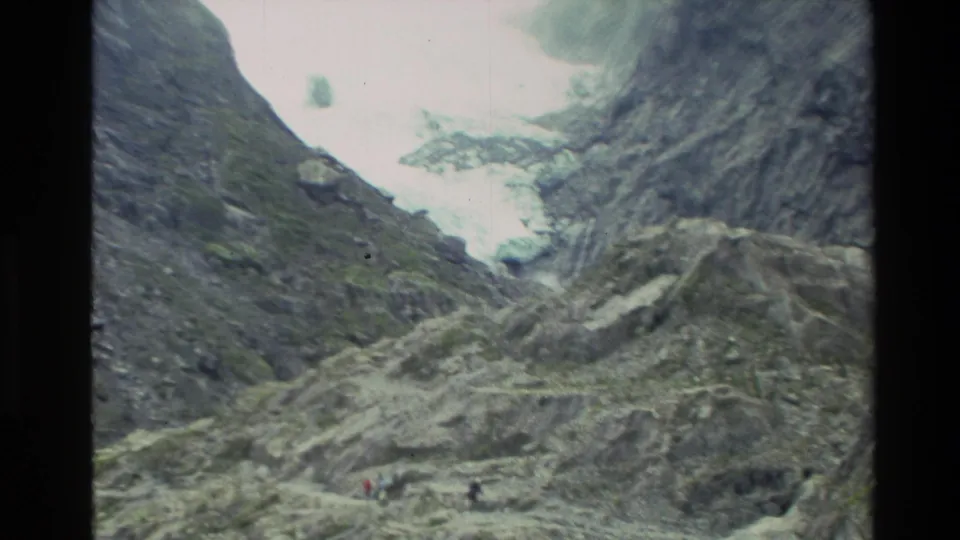 1982 New Zealand 8mm frame showing a glacier between steep rocky slopes, with tiny hikers on the moraine for scale.