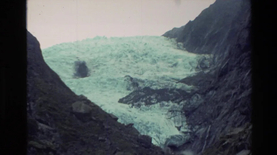 1982 New Zealand home-movie frame looking across pale glacier ice and dark alpine slopes from a rocky vantage point.
