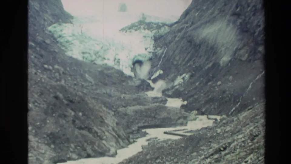 1982 New Zealand 8mm frame showing glacier ice above a rocky valley floor with a pale meltwater stream below.