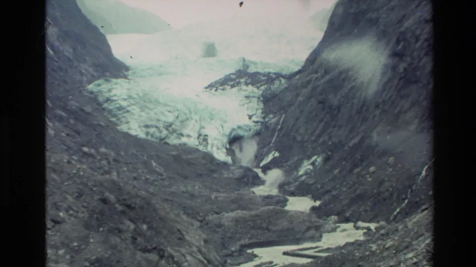 A cool-toned 8mm view of a New Zealand alpine valley, with glacier ice at the back and a stream winding through rock below.