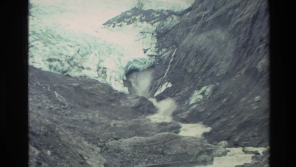 The 1982 New Zealand frame moves closer to the glacier outlet, showing pale ice, dark rock walls, and white water below.