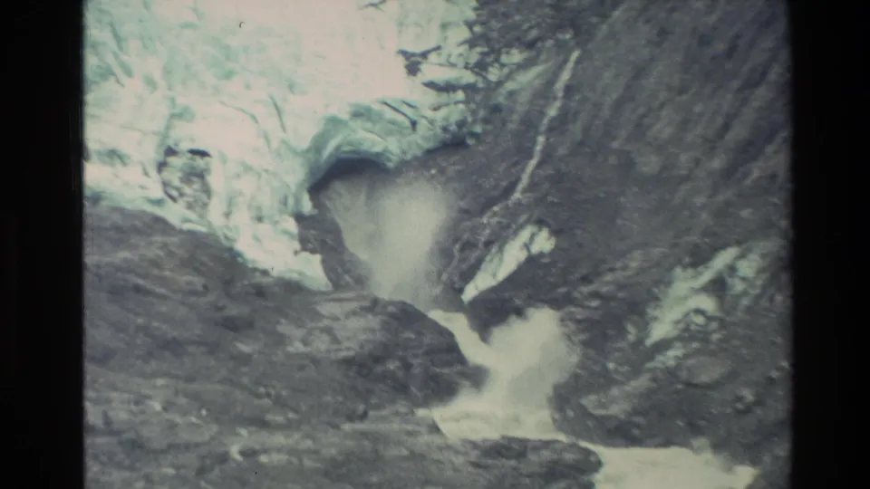 A tight 8mm frame of the New Zealand alpine scene shows pale glacier ice and rushing meltwater framed by dark rocky slopes.