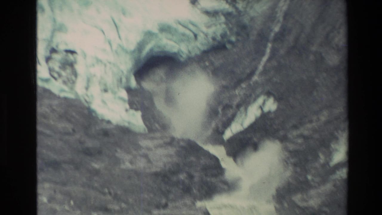 1982 New Zealand 8mm frame showing a white torrent emerging from a dark mountain ice-cave opening between snow, ice, and rock.