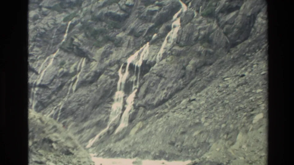 1982 New Zealand 8mm frame showing a bright narrow waterfall crossing a steep dark rock face with loose stones below.